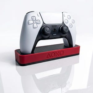Könnte beinhalten: Ein weiß-schwarzer PlayStation 5-Controller auf einer roten Basis. Auf der Basis steht der Text "ΔΟΧΟ". Der Controller hat ein modernes Design mit verschiedenen Tasten und Joysticks.