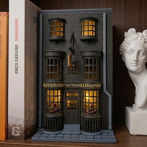 Może przedstawiać: Szczegółowy kącik książkowy z miniaturową repliką sklepu z różdżkami Olivandera z kolekcji Diagon Alley. Model ma ceglaną fasadę, podświetlane okna i nazwę sklepu. Jest wystawiony na półce z książkami i popiersiem.