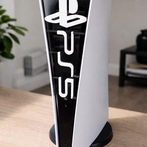 Könnte beinhalten: Eine weiße und schwarze PlayStation 5 Konsole steht auf einer Holzoberfläche. Die Konsole hat ein weißes Äußeres mit einem schwarzen Inneren und das PlayStation-Logo und "PS5" in Weiß.