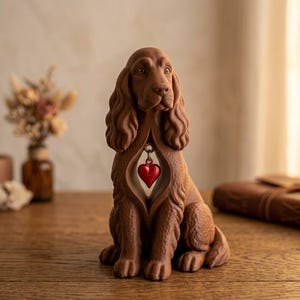 Op de afbeelding: Een bruin, getextureerd hondenbeeldje in een zittende houding. De hond heeft lange, golvende oren en een holle borstkas met daarin een kleine, glanzend rode hartvormige bedel die aan een zilverkleurige metalen ring is bevestigd.