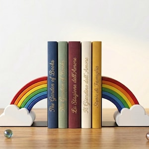Op de afbeelding: Een regenboogvormige boekensteunenset met witte wolkenbases ondersteunt een rij boeken. De boeken zijn in verschillende kleuren, met gouden letters die "The Garden of Books", "La Stagione dell'Amore", "Il Giardino dell'Amore" en "Storie di Famiglia" aangeven.