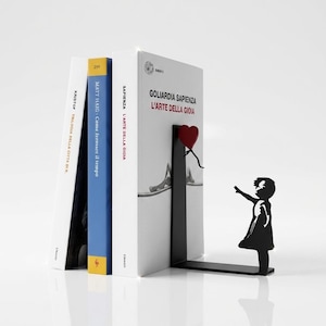 Può includere: Un fermalibri in metallo nero con la sagoma di un bambino che raggiunge un palloncino rosso a forma di cuore. Il fermalibri sostiene diversi libri, tra cui uno intitolato "GOLIARDIA SAPIENZA L'ARTE DELLA GIOIA".