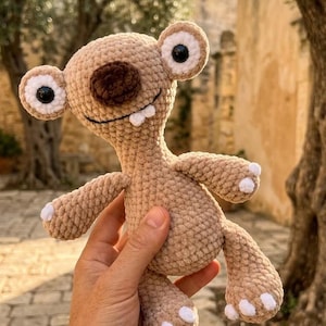 Peut inclure: Une peluche crochetée marron clair avec une grosse tête, deux grands yeux ronds, un nez marron foncé et deux petites dents blanches. La créature présente une surface en fil texturé, des bras et des jambes courts avec des pointes blanches.