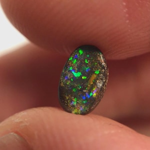 May include: A small, oval-shaped black opal gemstone with vibrant flashes of green, blue, and yellow. The opal is held between two fingers, showcasing its natural, polished surface and unique color play. The gemstone is a natural, untreated opal.