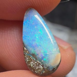 Könnte beinhalten: Ein tropfenförmiger Boulder-Opal-Edelstein mit einem lebendigen Spiel aus irisierenden Blau- und Grüntönen auf der polierten Oberfläche, kontrastiert durch eine texturierte, dunkelbraune Eisensteinmatrix an der Basis des Steins.