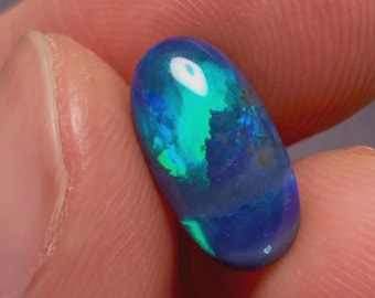 May include: An oval-shaped opal gemstone, held between fingers, displays a vibrant play of color. The stone's surface reflects shades of blue, green, and hints of white, creating a mesmerizing display. The opal's polished surface catches the light, showcasing its unique internal patterns.