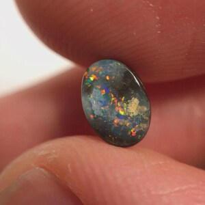 May include: An oval black opal gemstone, displaying a spectrum of colours such as red, orange, yellow, and blue. The opal is held between fingers, highlighting its polished surface and the unique play-of-colour effect.