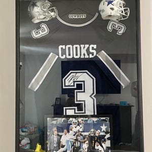 Könnte beinhalten: Gerahmte Präsentation von Dallas Cowboys-Erinnerungsstücken. Sie enthält ein marineblaues Trikot mit dem Namen "Cooks" und der Nummer 3, zwei Helme, Sammelkarten und ein Foto. Der Rahmen ist schwarz.