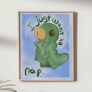 May include: A framed art print featuring a green cartoon caterpillar with a tan head, resting on a light blue background. The text "I just want to nap" is written in green. The artwork is in a light wooden frame.