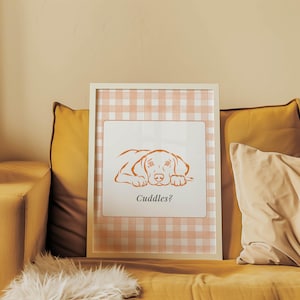 May include: A rectangular white picture frame containing a print with a light orange and white gingham background. The print features an orange line drawing of a dog lying down with the text Cuddles? printed in black below the illustration.