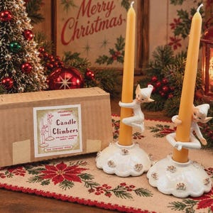 May include: Two white ceramic candle holders featuring elf-like figures climbing tall yellow taper candles. The base of each holder is round with iridescent accents. A cardboard box is included with a label reading Decorative Ceramic Candle Climbers.