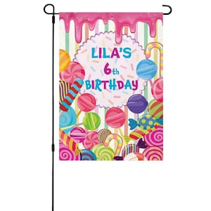 May include: A colorful garden flag with a candy theme. The flag features the text "Lila's 6th Birthday" in a bubble font, surrounded by various candies, lollipops, and candy canes. The top of the flag has a pink frosting design with sprinkles.