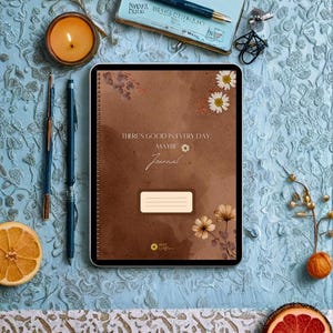 Puede incluir: Un diario de espiral marrón con el texto "THERE'S GOOD IN EVERY DAY. MAYBE Journal" e ilustraciones florales. El diario presenta una etiqueta rectangular, un pequeño icono de margarita y el nombre de la marca "ALLY WILDFLOWER" abajo.