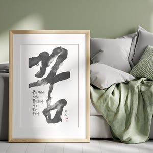 May include: A framed wall art print featuring large, bold black ink calligraphy brushstrokes on a white background. Smaller Korean text is printed to the left of the main character, with a small red stamp signature in the bottom right corner.