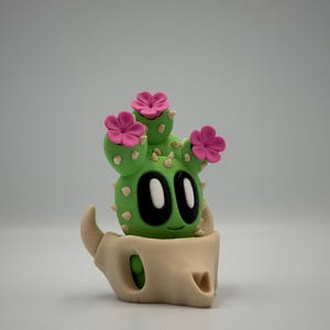 May include: A whimsical green cactus figurine with pink flower accents and a smiling face. The cactus is nestled in a tan, skull-shaped pot. The figurine is a unique decorative item.