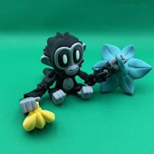 May include: A whimsical figurine of a cartoon monkey in black and gray, holding a bunch of yellow bananas and a light blue flower. The monkey has large, round eyes and is set against a solid green background.