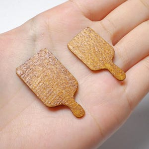 May include: Two miniature wooden chopping boards, each with a handle. The boards are rectangular with a textured surface and a light brown hue. They are shown on a hand, possibly for crafts or decorative use.