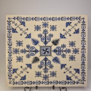 May include: A square, off-white embroidered textile with a symmetrical blue and black floral design. The central motif is a stylized cross, surrounded by various floral and geometric patterns. The border features a repeating leaf pattern.