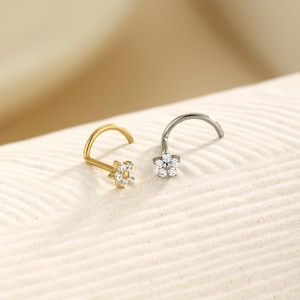 May include: Two nose rings with flower designs. One is gold-colored with clear gemstones, and the other is silver-colored with clear gemstones. Both have a curved post for insertion.