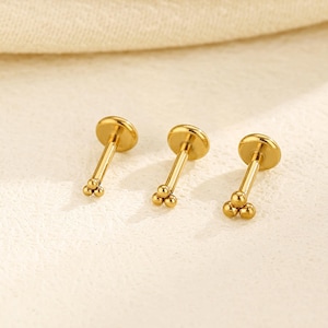May include: Three gold-colored labret studs. Each stud features a flat, round top and a straight post. The bottom of each post has three small, round beads. The studs are displayed on a light-colored, textured surface.