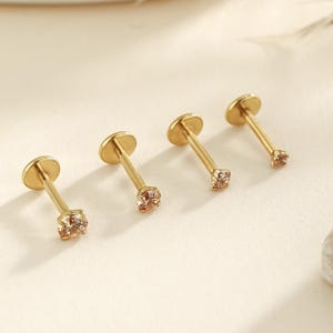 May include: Four gold-colored, flat-backed earrings with a small, square, peach-colored gemstone at the end of each post. The earrings are arranged in a row, with the gemstones facing the viewer. The earrings are on a light-colored surface.