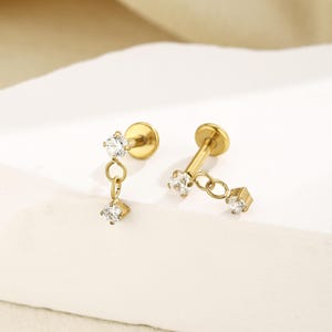 May include: A pair of gold-tone flat back stud earrings featuring a small round clear gemstone at the post and a dangling square clear gemstone charm attached by a circular link. The earrings have a polished metallic finish and a secure backing.