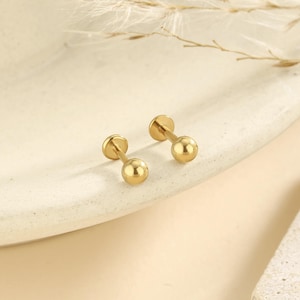 May include: A pair of gold-colored stud earrings. Each earring features a small, round ball at the end of a post, with a flat disc on the other end. The earrings are displayed on a light-colored surface.