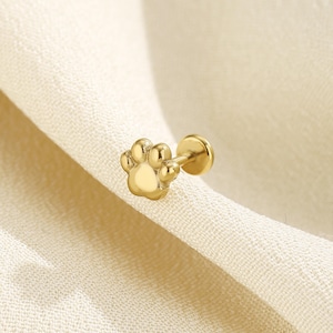May include: A gold-toned stud earring featuring a paw print design with a flat circular back post. The earring has a polished metallic finish and is shaped like a small animal paw with four distinct rounded toes and a central pad.