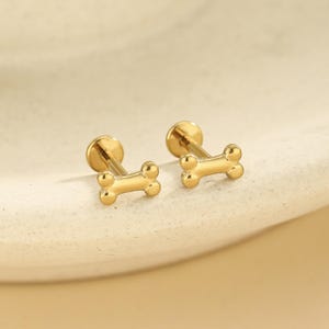 May include: A pair of gold-toned stud earrings, each featuring a polished dog bone shape. The earrings have a flat circular backing and a post, showcasing a smooth, reflective metallic finish that catches the light with a bright yellow hue.