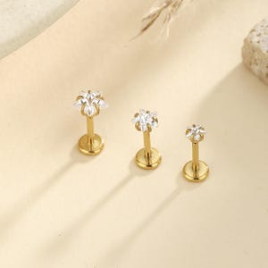 Könnte beinhalten: Drei goldfarbene Lippen- oder Ohrstecker mit sternförmigen, klaren Kristallaufsätzen. Die Stecker haben eine flache, kreisförmige Basis und einen schlanken Stab. Die Kristalle funkeln vor dem warmen, neutralen Hintergrund.