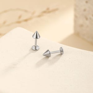 May include: Two silver-colored, cone-shaped stud earrings. Each earring has a pointed cone top and a straight post with a flat disc backing. The earrings are displayed on a light-colored surface, with one standing upright and the other lying horizontally.