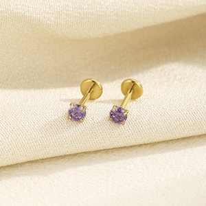 May include: A pair of gold-colored stud earrings with a small, round, purple gemstone. The earrings have a flat, circular backing and a slender post. The earrings are displayed on a cream-colored fabric.
