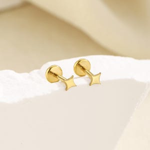 May include: A pair of gold-toned stud earrings featuring a four-pointed star design. Each earring consists of a flat, circular back piece connected to a thin post that leads to a small, polished star-shaped front element with curved edges.