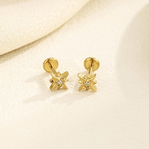 May include: A pair of gold-coloured star-shaped stud earrings. Each earring has a small, clear gemstone at the centre of the star design. The earrings are displayed on a light-coloured surface, with a soft, neutral background.