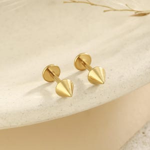 May include: Two gold-colored stud earrings. Each earring features a flat, round backing and a conical, pointed front. The earrings are made of a shiny, metallic material and are displayed on a neutral-toned surface. The earrings are designed for ear piercings.