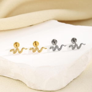 May include: Close-up of four stud earrings with a snake design. Two are gold-colored, and two are silver-colored. The earrings are on a white, textured surface with a soft, neutral background.