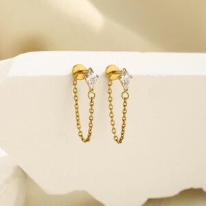 May include: A pair of gold-toned stud earrings featuring a kite-shaped clear gemstone attached to a delicate gold chain that loops back to the post. The earrings have a flat circular backing and a polished metallic finish throughout the design.