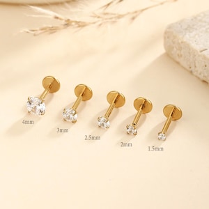 May include: Five gold-colored labret studs with clear, round cubic zirconia stones. The sizes are 4mm, 3mm, 2.5mm, 2mm, and 1.5mm. The studs have a flat disc backing.