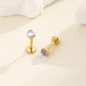 Könnte beinhalten: Ein Paar flache Labret-Stecker in Goldoptik mit einem kleinen, runden, halbtransparenten blauen Cabochon-Stein an der Oberseite. Jedes Stück besteht aus einem geraden Stift, einer runden Basis und einer glatten, polierten Metalloberfläche.