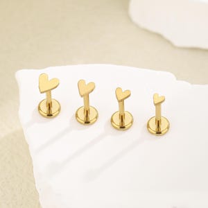 Könnte beinhalten: Vier goldfarbene Ohr- oder Lippenstecker in Herzform. Jeder Stecker hat ein herzförmiges Oberteil, einen geraden Stab und einen runden Sockel. Die Stecker sind in einer Reihe angeordnet, wobei die Herzen nach vorne zeigen. Der Hintergrund ist eine neutrale Farbe.