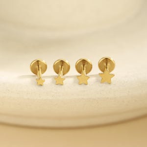 May include: Four gold-toned flat back stud earrings arranged in a row, each featuring a five-pointed star design. The stars increase in size from left to right, and each earring has a circular flat base attached to a thin post.