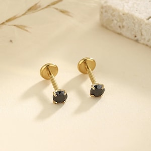May include: Two gold-colored earrings with a small, round, black gemstone at the base of each post. The earrings have a flat, circular top and a slender post. The background is a soft, neutral color.