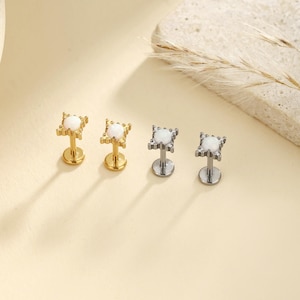 May include: Four stud earrings. Two are gold-toned, and two are silver-toned. Each earring features a square-shaped design with a central white stone. The earrings are displayed on a light-colored surface.