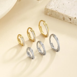 May include: Six hoop earrings in gold and silver tones, each adorned with small, sparkling stones. The earrings are arranged in a group, showcasing their design and variations. The background is a neutral beige.