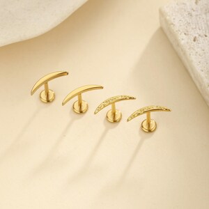 May include: Four gold-colored crescent moon-shaped stud earrings. Two earrings are plain, while the other two have small, sparkling embellishments. The earrings are displayed on a light beige surface.