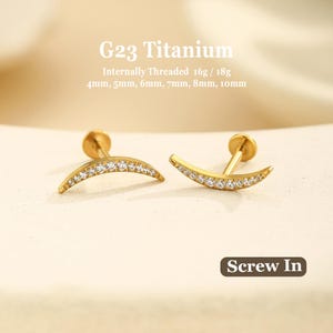May include: Two gold-tone G23 titanium stud earrings featuring a curved crescent moon shape encrusted with small clear stones. The text reads G23 Titanium, Internally Threaded 16g / 18g, 4mm, 5mm, 6mm, 7mm, 8mm, 10mm, and Screw In.