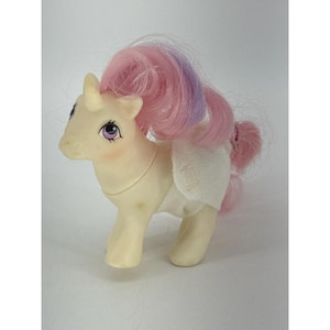 May include: A cream-colored unicorn toy with a small horn on its forehead. The figure has purple eyes, pink blush on its cheeks, and a long, flowing mane and tail featuring shades of pink and purple. It wears a simple white fabric diaper.