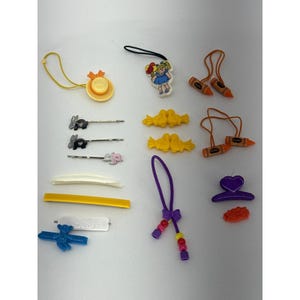 Puede incluir: Una colección de accesorios para el cabello que incluye un dije de sombrero amarillo, un dije de personaje, ligas para el cabello con forma de crayón, pasadores de conejo y animales, un peine blanco, una hebilla amarilla y un clip de oso azul.