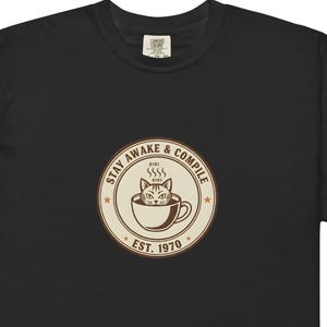 Puede incluir: Camiseta negra con un gráfico circular que presenta un gato en una taza de café, vapor y el texto "STAY AWAKE & COMPILE" y "EST. 1970". La camiseta tiene una etiqueta Comfort Colors.