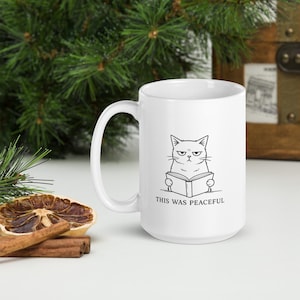 Könnte beinhalten: Weiße Keramiktasse mit einer Schwarz-Weiß-Illustration einer grummeligen Katze, die ein Buch liest. Der Text "THIS WAS PEACEFUL" steht unter der Katze. Die Tasse steht auf einer weißen Oberfläche.